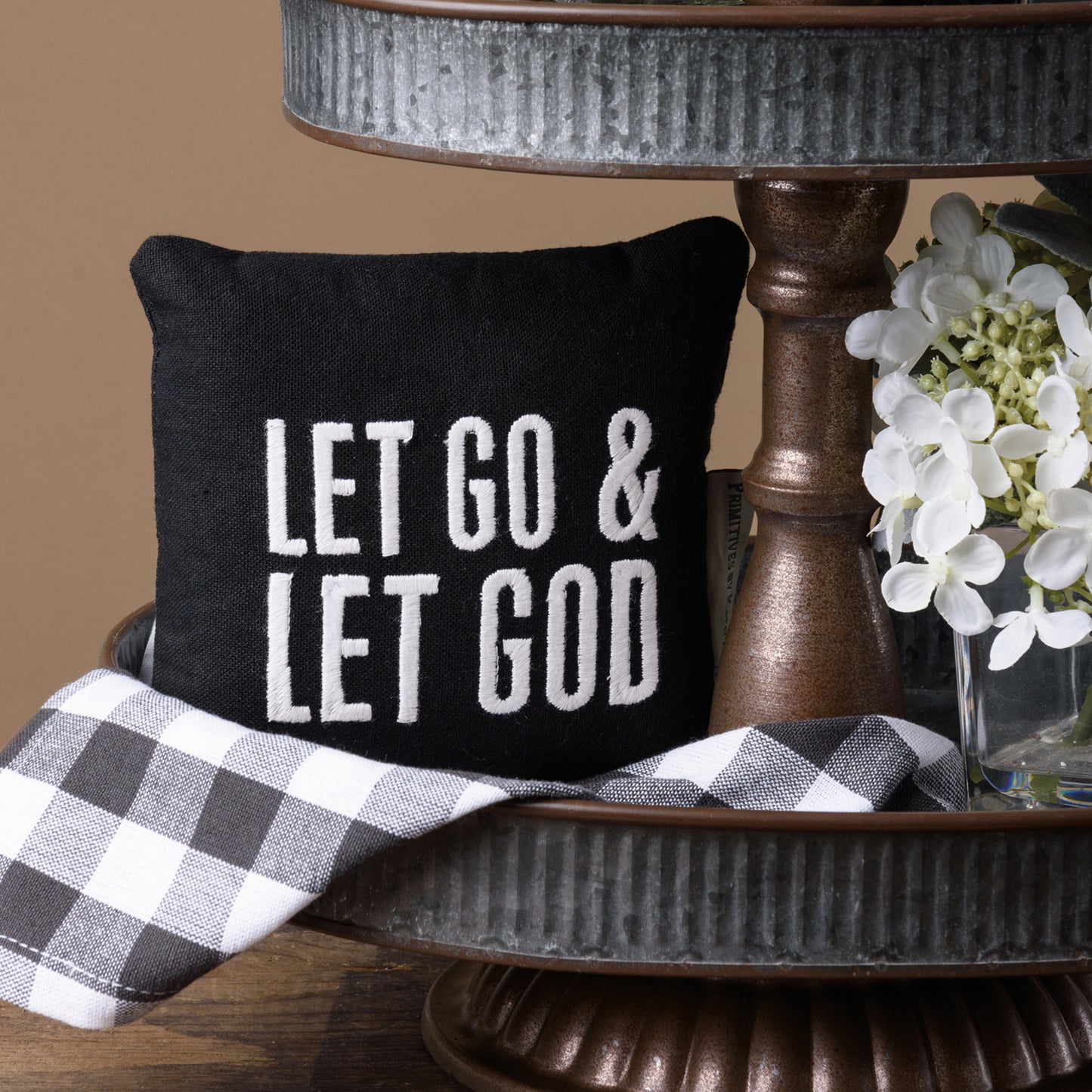 a small black pillow that reads Let Go & Let God sits on the bottom tray of a metal tiered tray with a gray and white checked towel under the pillow and a small white flower on the right