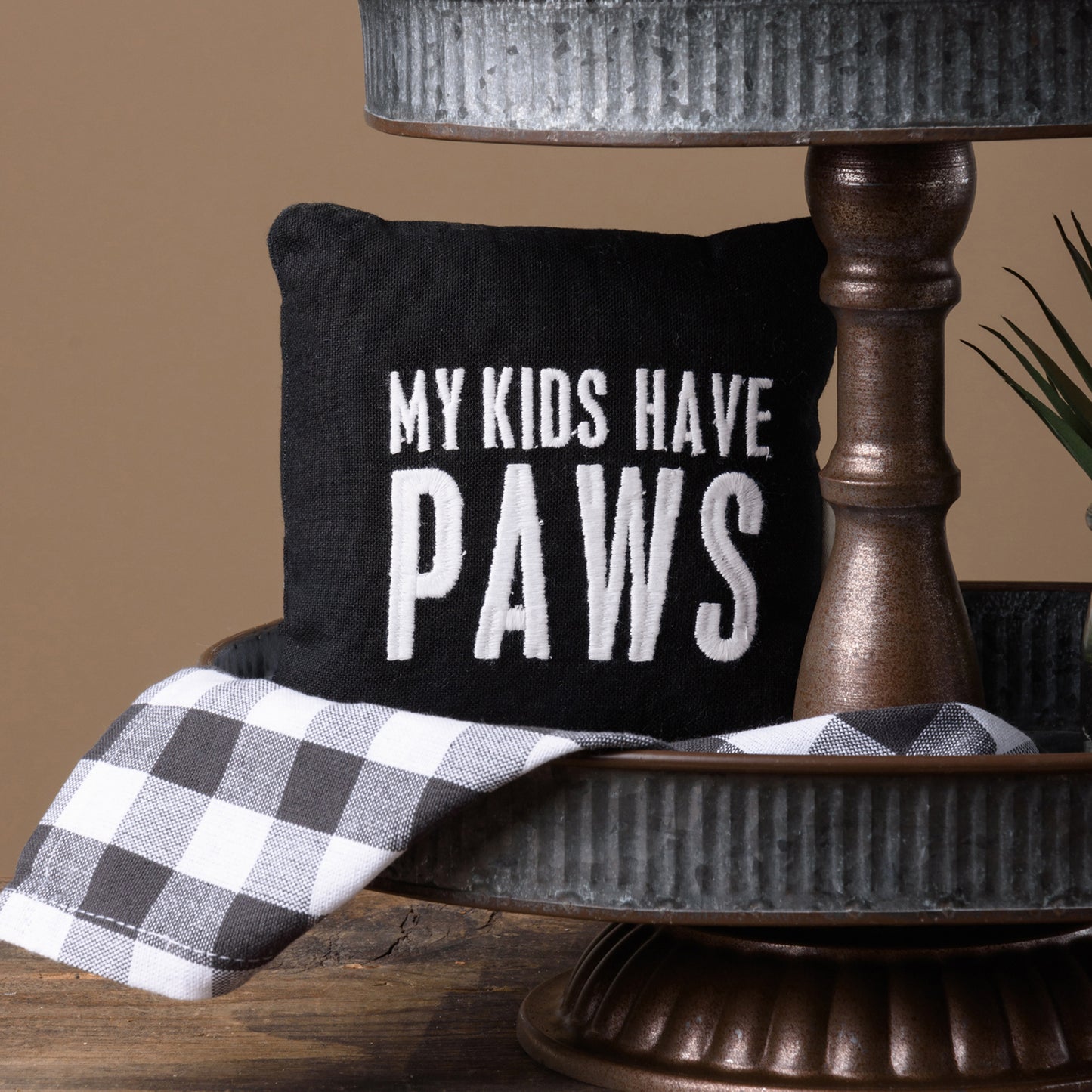 a small black and white pillow that says my kids have paws sits on the bottom tray of a tiered stand, with a gray and white check towel under the pillow