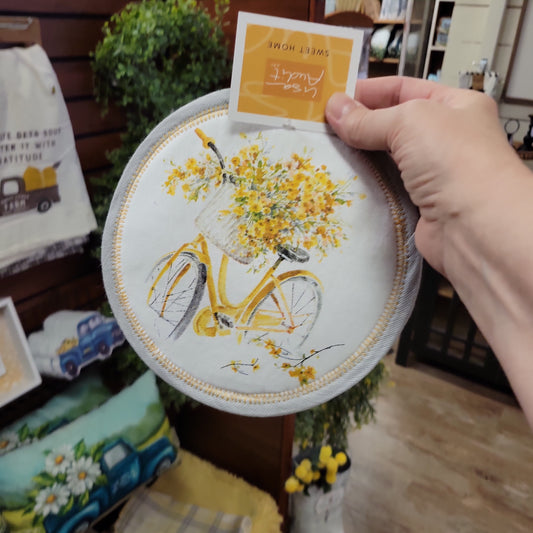 round white potholder with gray trim and yellow stitching featuring a yellow bicycle in the middle with cascading yellow flowers 