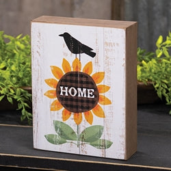 distressed block with a sunflower that says HOME and a black crow on top, sitting on a black shelf with greenery